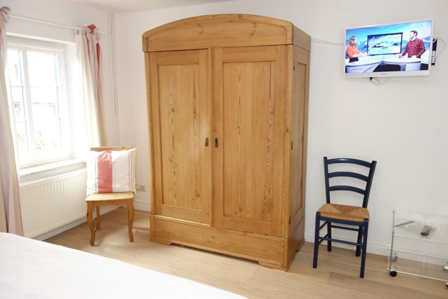 Ein Zimmer mit einem Holzschrank, einem kleinen Holzstuhl, einem an der Wand befestigten Fernseher und einem Fenster mit Vorhängen.