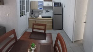 Eine kompakte Küche mit Kühlschrank, Spüle und Holzschränken. Ein Esstisch mit Stühlen und ein kleiner Topfpflanze sind ebenfalls vorhanden.