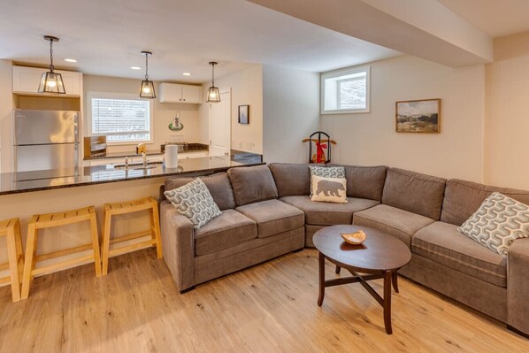 A modern living room with a sectional sofa, wooden stools, a round coffee table, and a kitchen area with a refrigerator, sink, and cabinets.