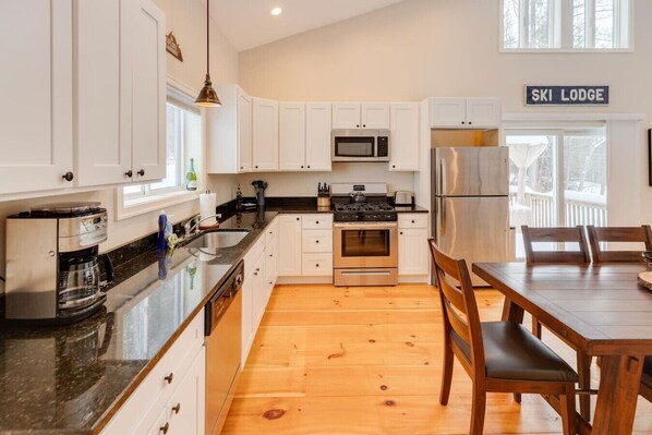 A modern kitchen with white cabinets, a stainless steel refrigerator, a microwave, and a stove. There is a wooden dining table with chairs and a sign that reads "SKI LODGE". - Spacious House in the White Mountains! (Thornton)