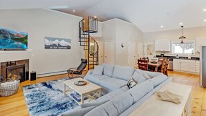 Ein modernes Wohnzimmer mit Ecksofa, Kamin, Fernseher und einer Küche im Hintergrund.