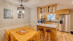 A modern kitchen with wooden cabinets, a stainless steel refrigerator, a dining table with chairs, and a hanging light fixture. - Loon Condo with Fireplace (Woodstock)