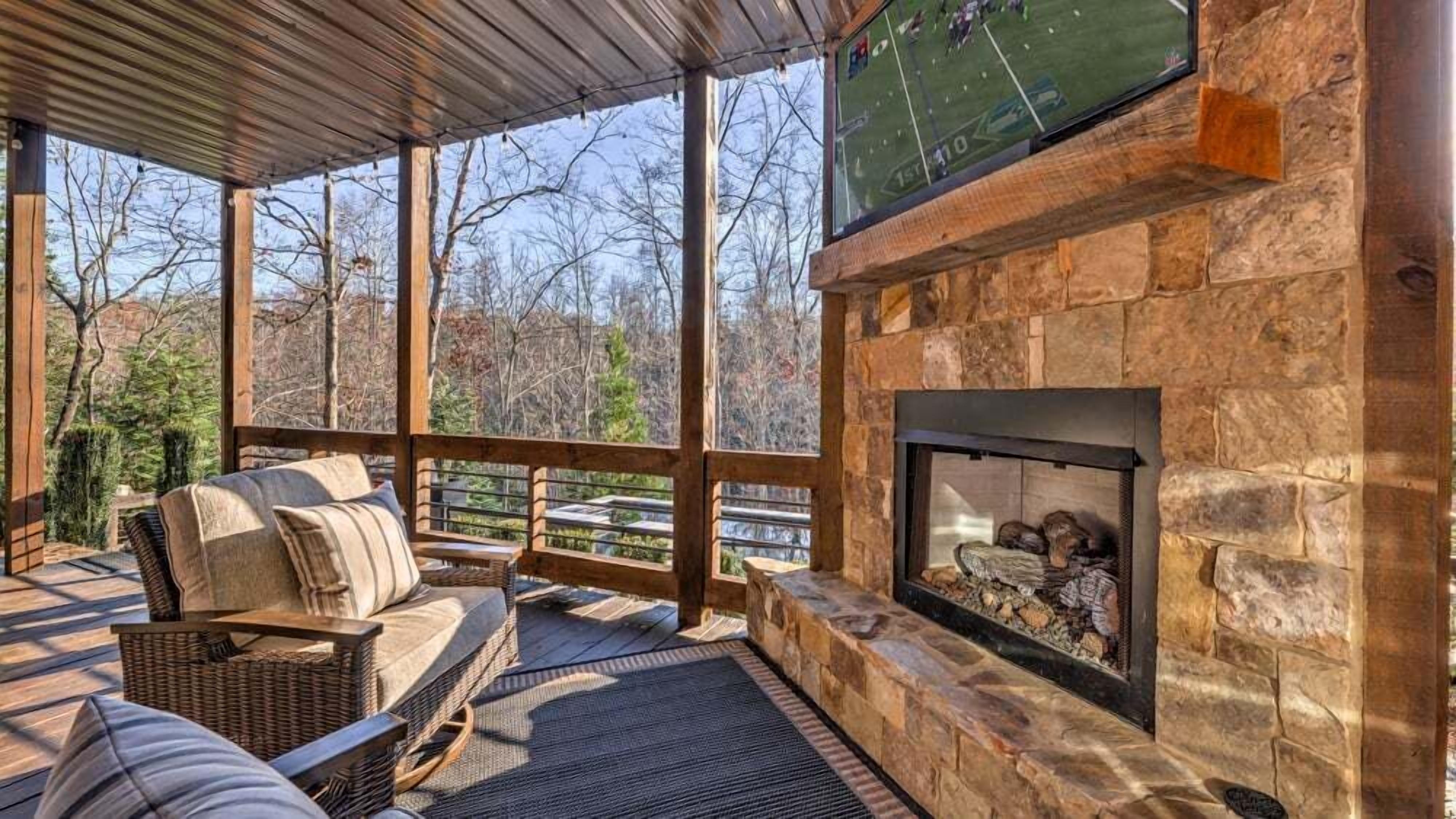 A covered deck with a stone fireplace, outdoor seating, and a large window overlooking a sports field.