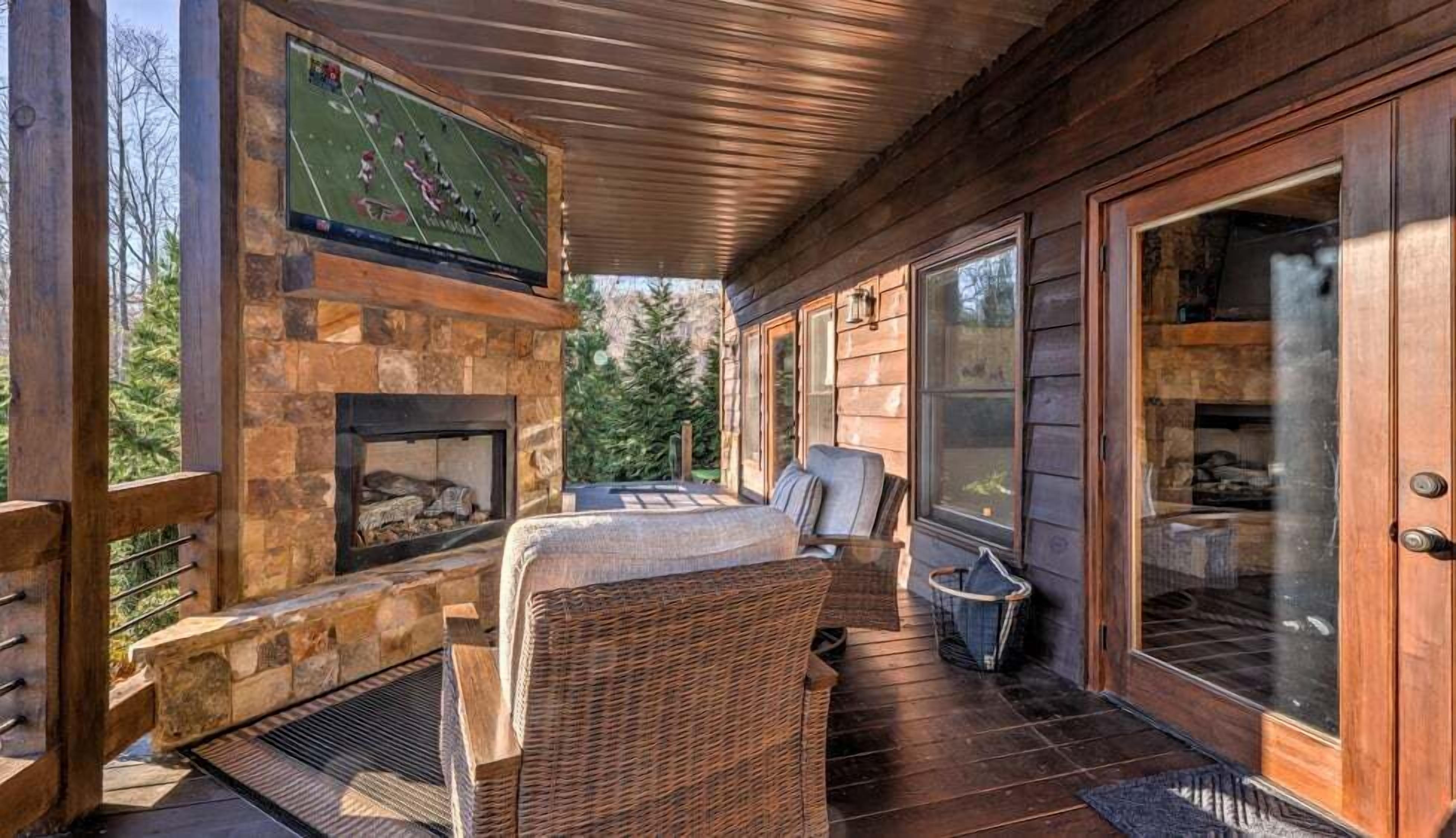 A covered outdoor seating area with a stone fireplace, wicker furniture, and a television screen displaying a football game.