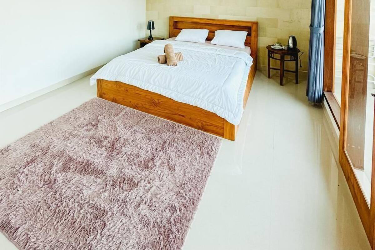 A bedroom with a wooden bed, a fluffy rug, a nightstand, a lamp, and an air conditioning unit on the wall.