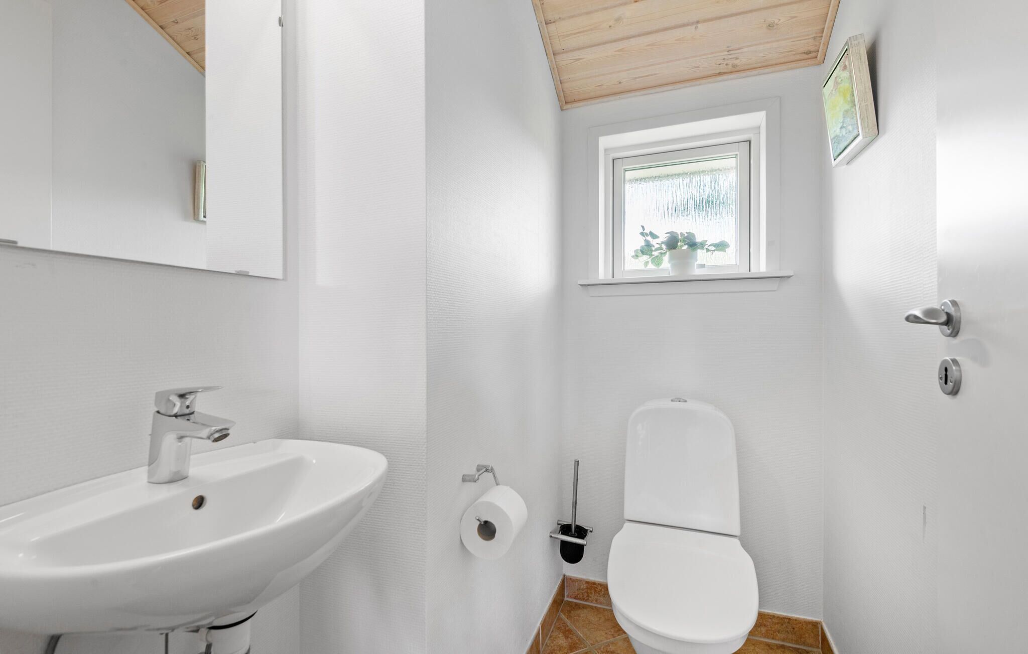 A small, white bathroom with a wooden ceiling, a white sink, a toilet, and a window with a view of greenery.