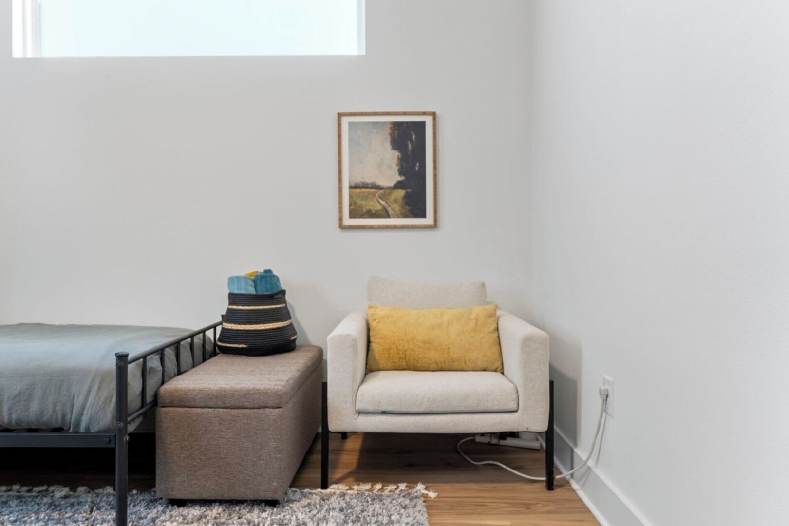 A small, modern bedroom with a bed, a grey ottoman, a white armchair, and a framed picture on the wall.