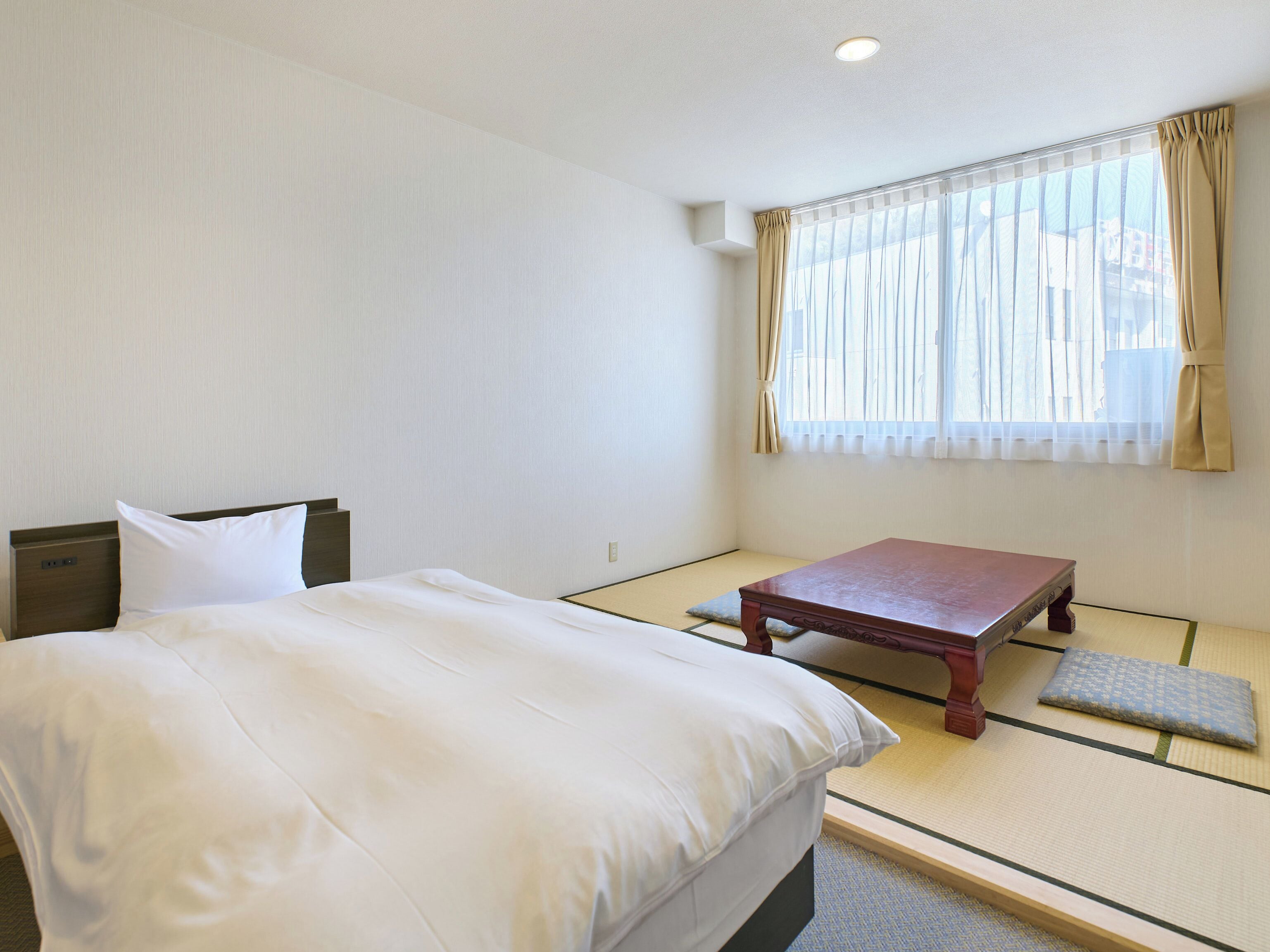 A hotel room with a bed, a wooden table, tatami flooring, and a window with curtains.