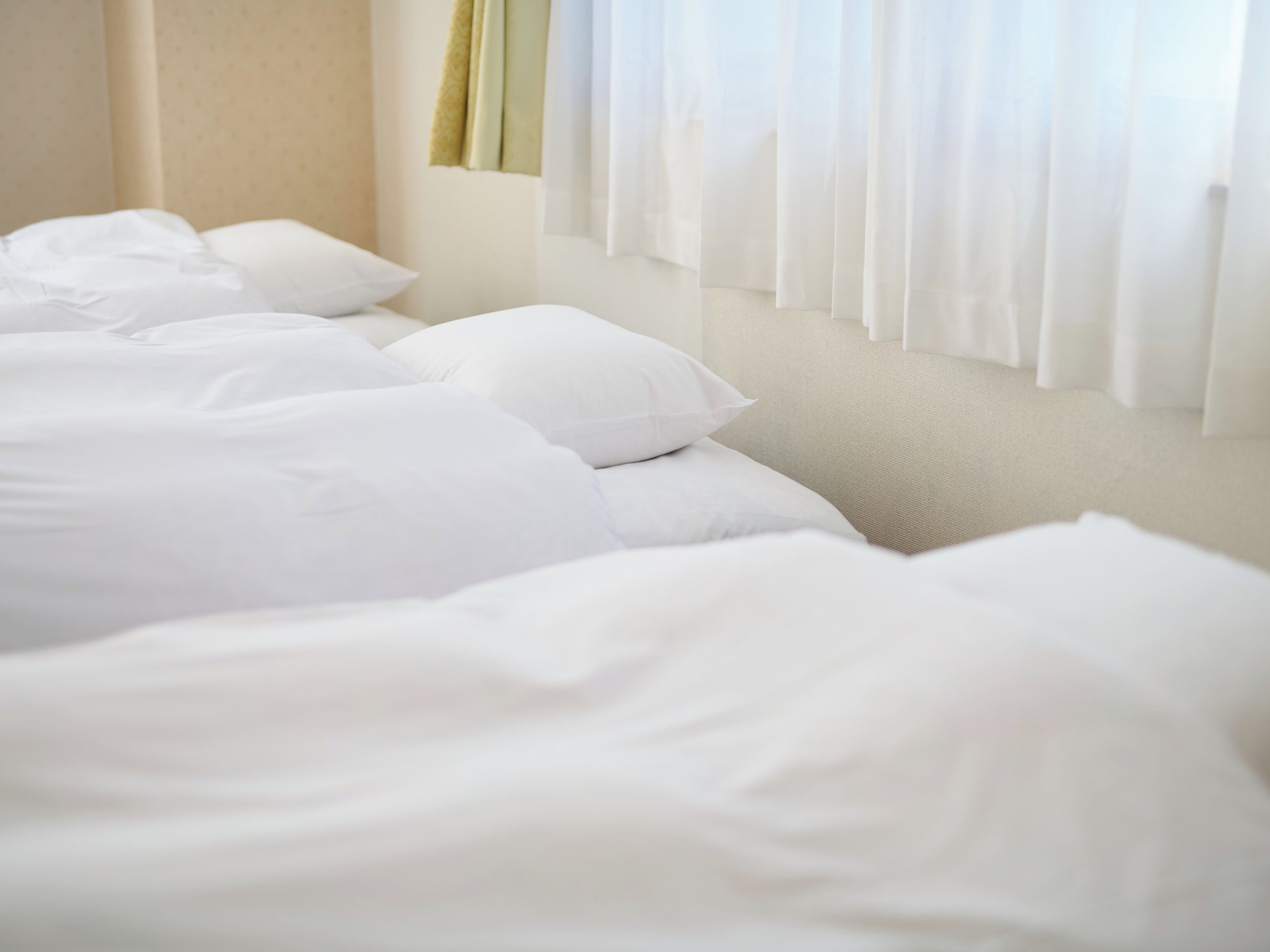 Een net opgemaakt bed met witte lakens en kussens in een kamer met een beige wand en een raam met witte gordijnen.
