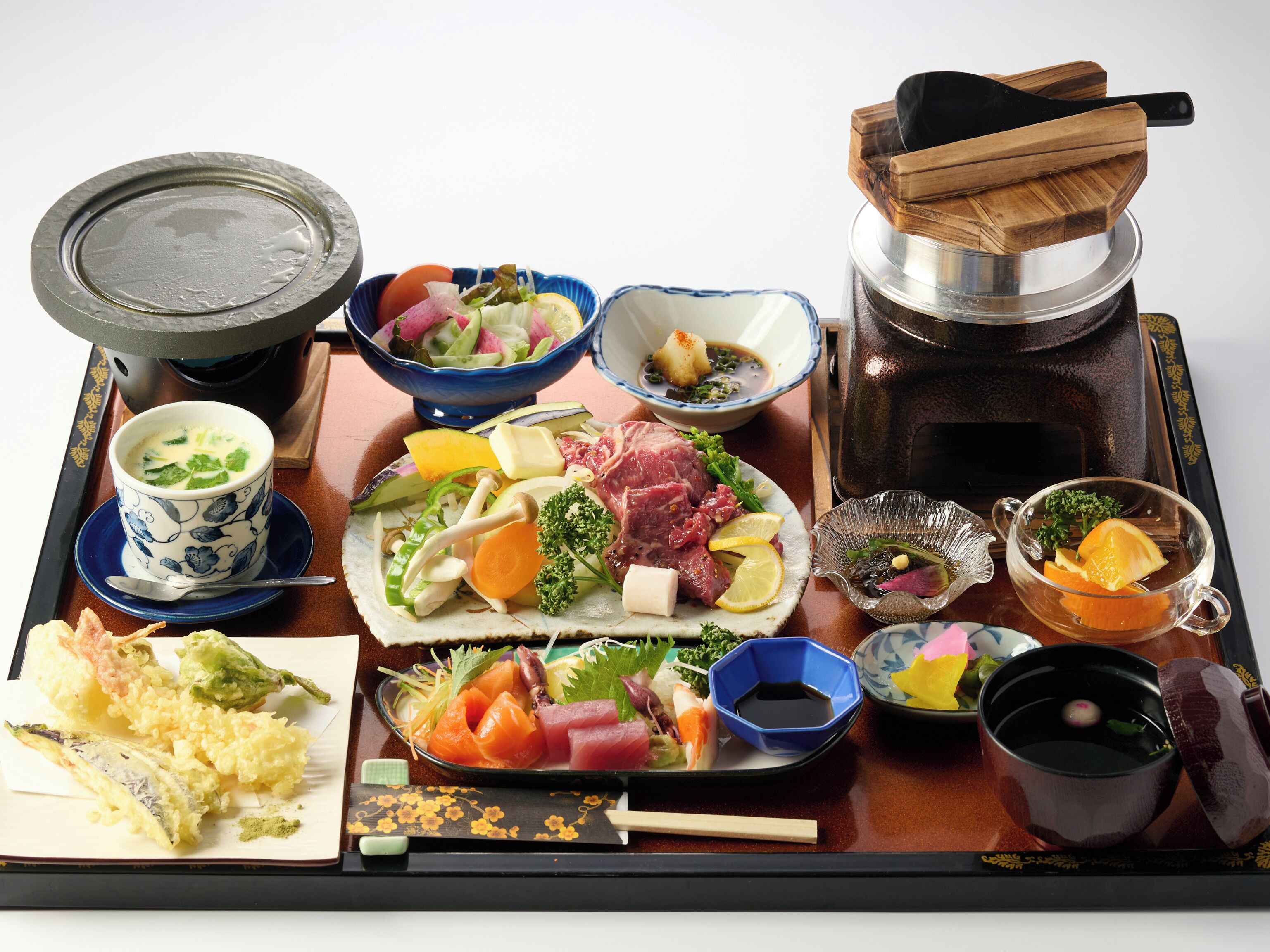 Un plato tradicional japonés de olla caliente con diversas verduras, carnes y mariscos, servido en una bandeja de madera con palillos y un juego de té.