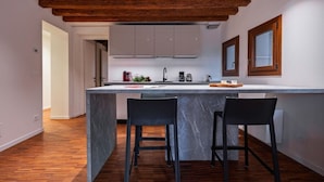 A modern kitchen with a marble island, wooden ceiling beams, and two black bar stools.