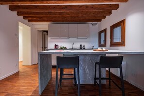 A modern kitchen with a marble island, wooden ceiling beams, and two black bar stools.