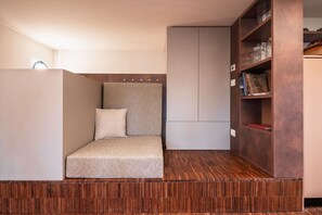 A compact room with a built-in sofa, a wooden floor, a wardrobe, and a shelf with books and decorative items.