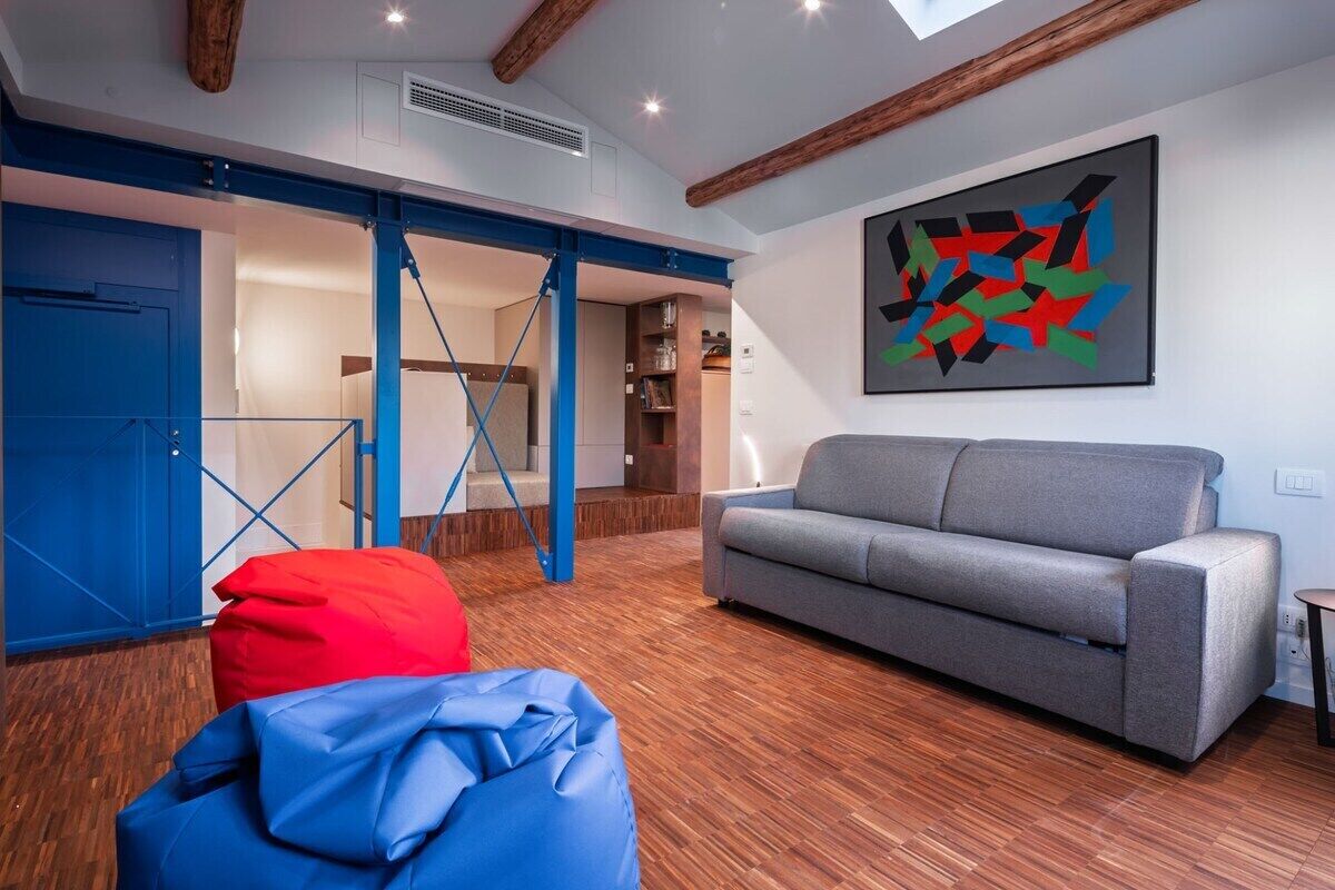Un salon moderne avec un canapé gris, des poufs rouges et bleus, et un tableau abstrait aux couleurs vives accroché au mur.