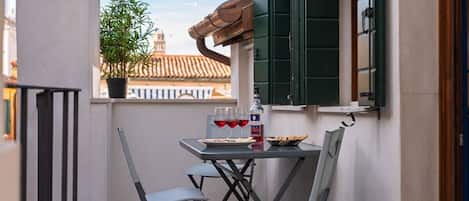 A balcony with a table, chairs, a potted plant, and a window with green shutters.