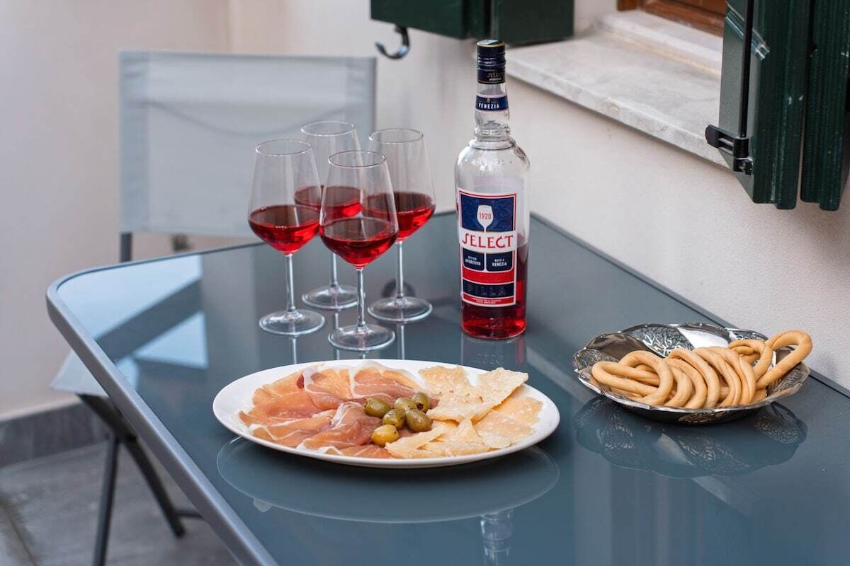 Een tafel met een fles drank, glazen rode wijn, een bord met vleeswaren, olijven en crackers.
