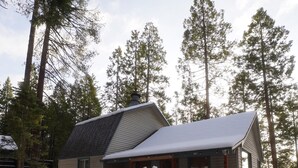 Una cabaña con terraza de madera y suelo cubierto de nieve, rodeada de altos pinos.
