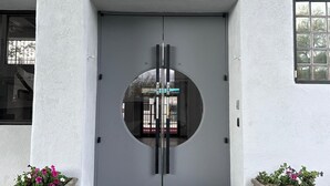 Een moderne entree met een grote, ronde glazen deur, aan weerszijden twee potplanten, en een betonnen vloer.