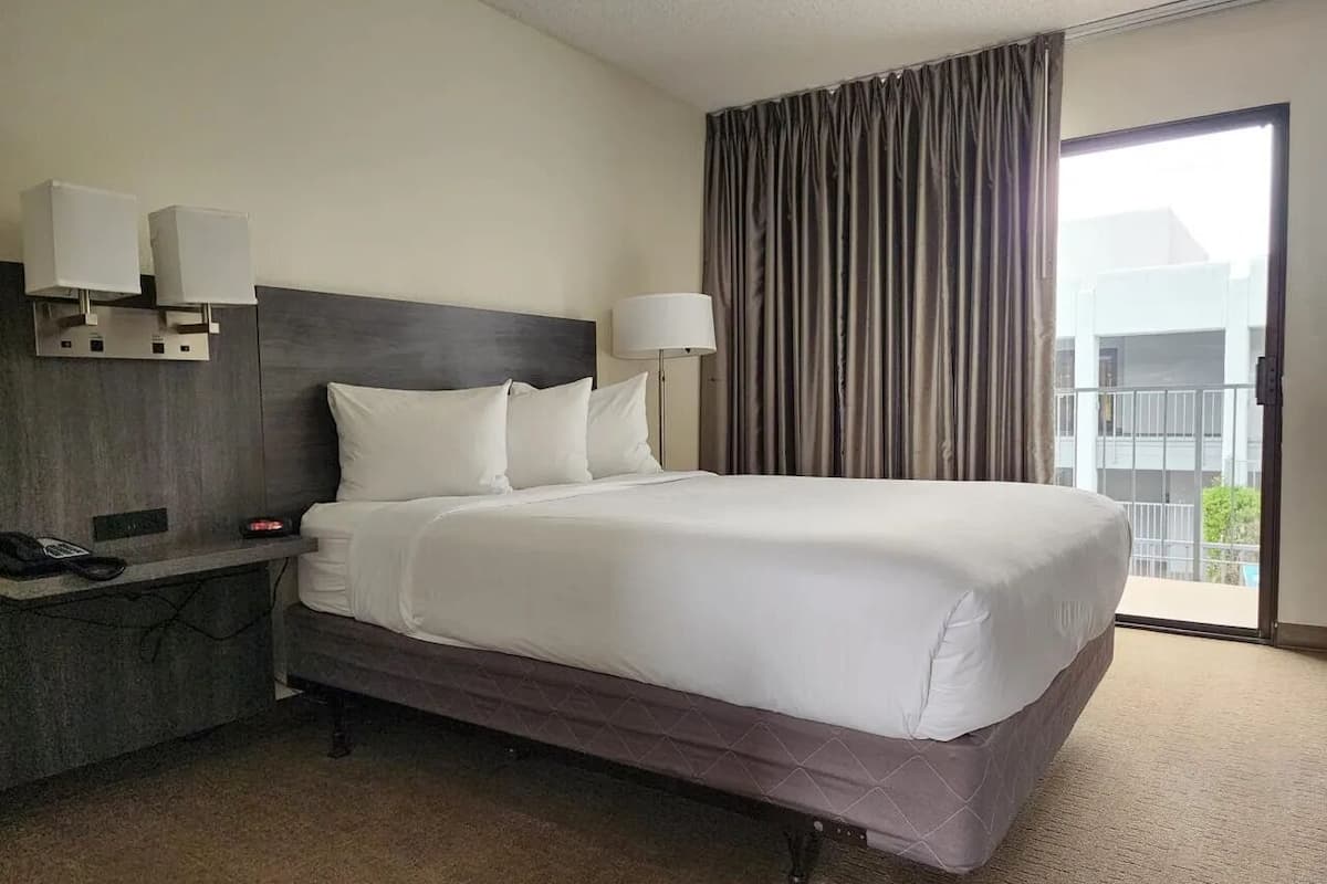 Habitación de hotel con una cama grande, una mesita de noche con lámpara, teléfono y vistas al exterior por una ventana.