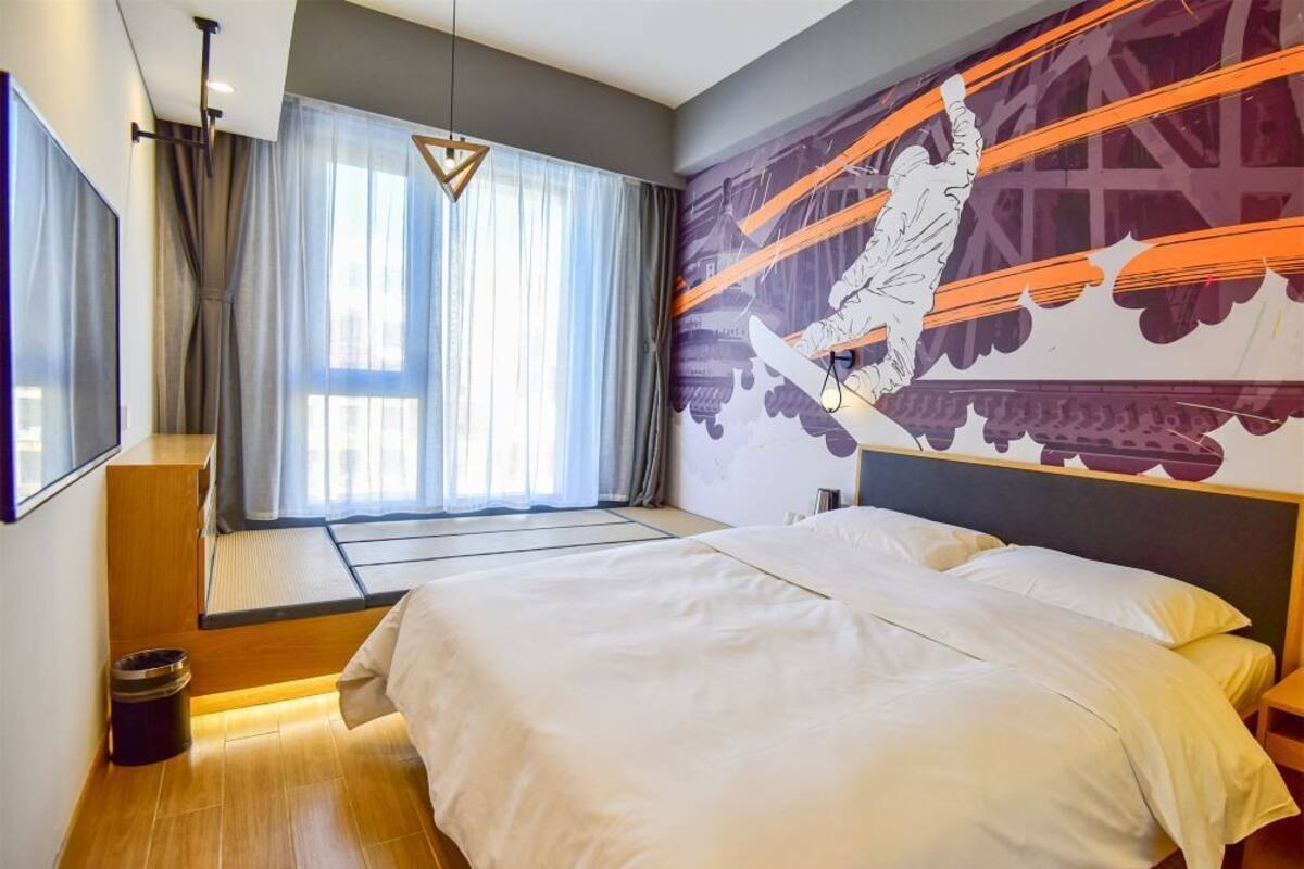Et moderne hotelværelse med en stor seng, et vindue med lette gardiner, et vægmaleri og et lille siddeområde med udsigt over byen.