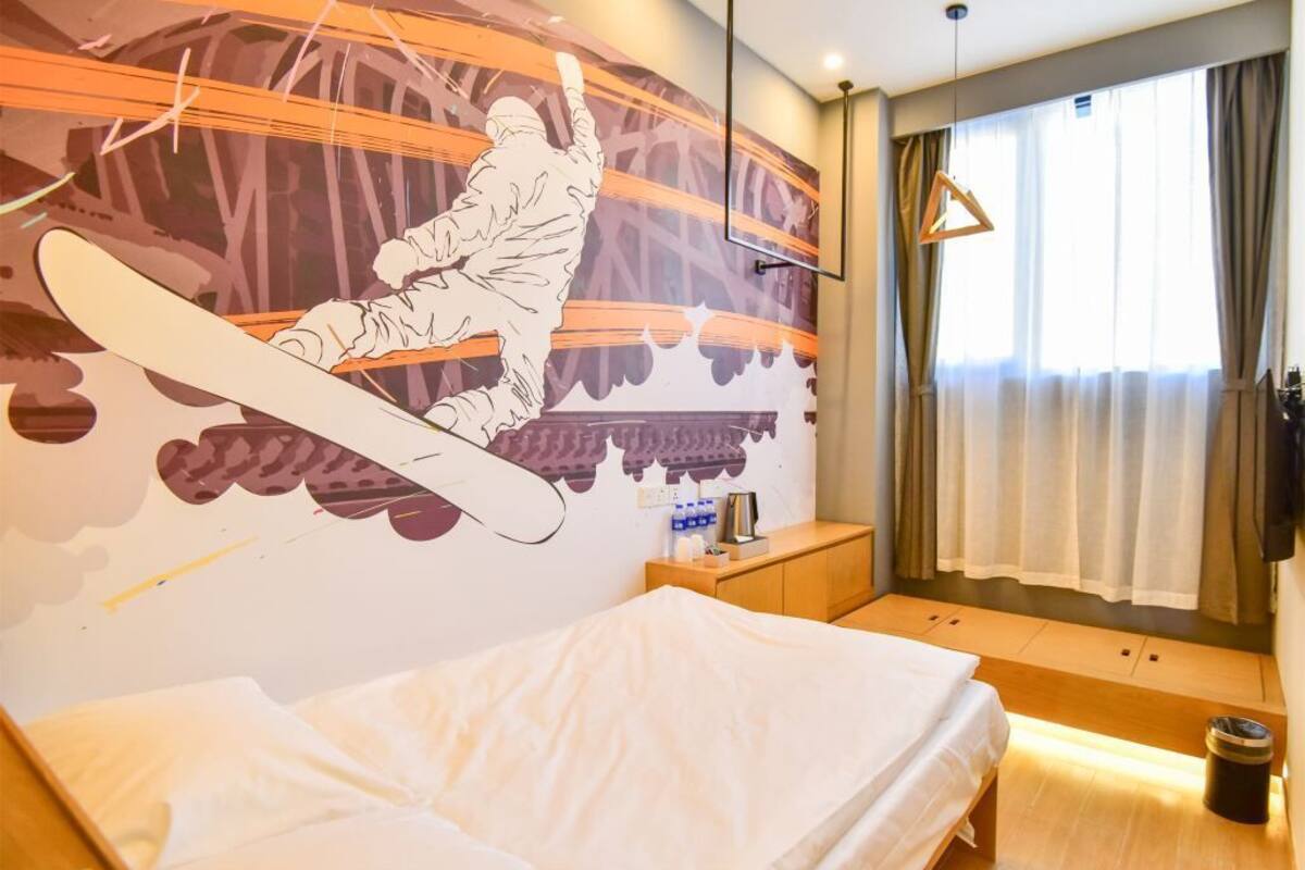 Et hotelværelse med seng, et natbord, et vægmaleri af en snowboarder, et vindue med gardiner og en hængende lysarmatur.