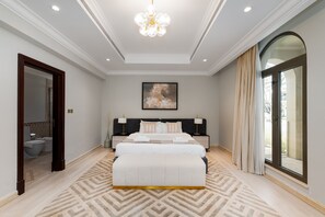 A spacious bedroom with a large bed, a nightstand, a bedside table, a chandelier, a painting on the wall, and a view through a large window.