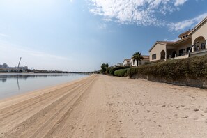 Ein Strand mit sandigem Ufer, ruhigem Meer und einer Reihe von Häusern mit Rundbogenfenstern und einem Ziegeldach.