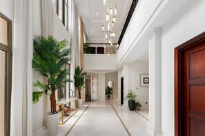 A spacious, well-lit hallway with a marble floor, large potted plants, and a wooden door on the right.
