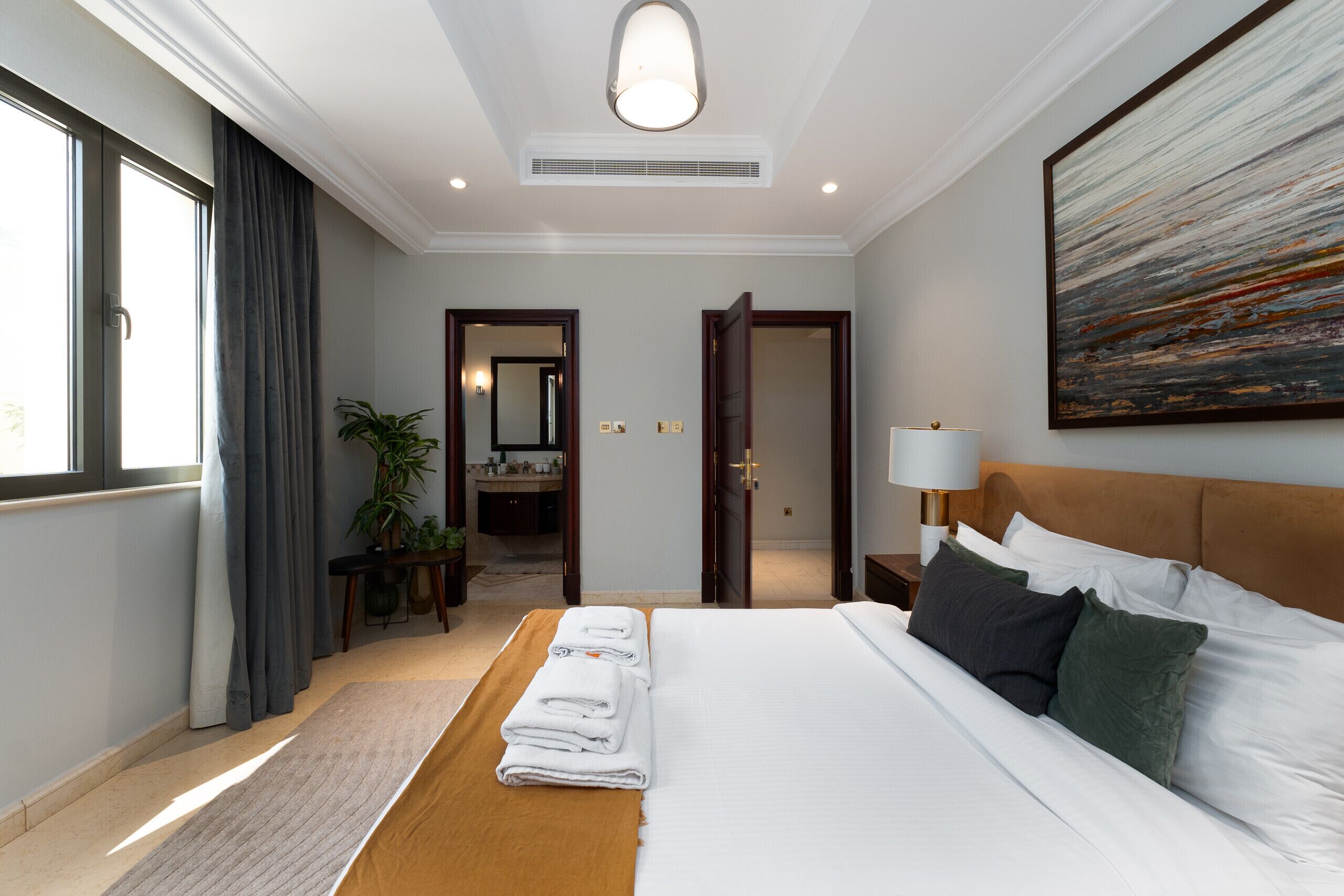 A hotel room with a large bed, a bedside table with a lamp, a painting on the wall, and a view of the bathroom through an open door.