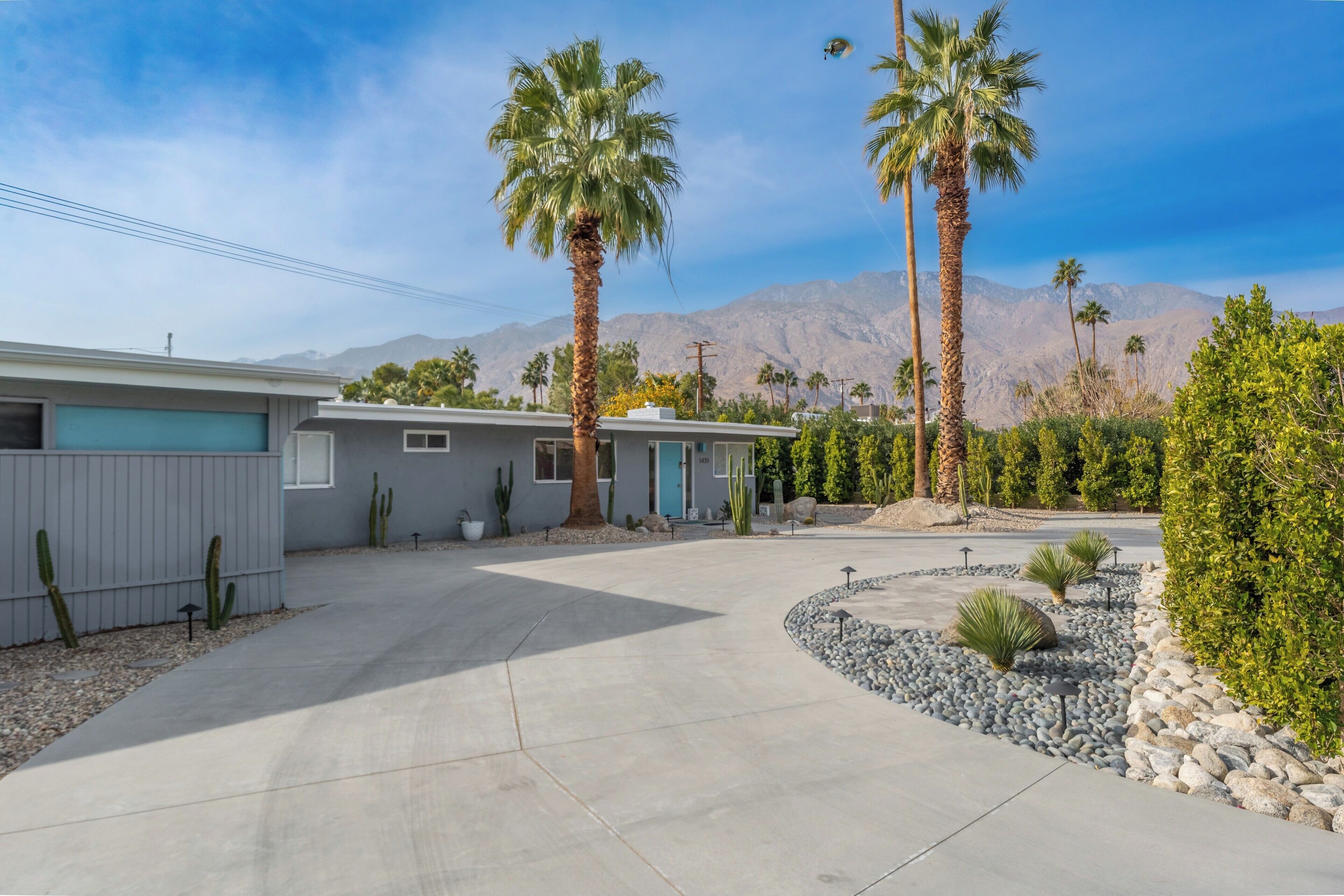 A modern two-story house with a driveway leading to a garage, surrounded by cacti and palm trees, with mountains in the background.
