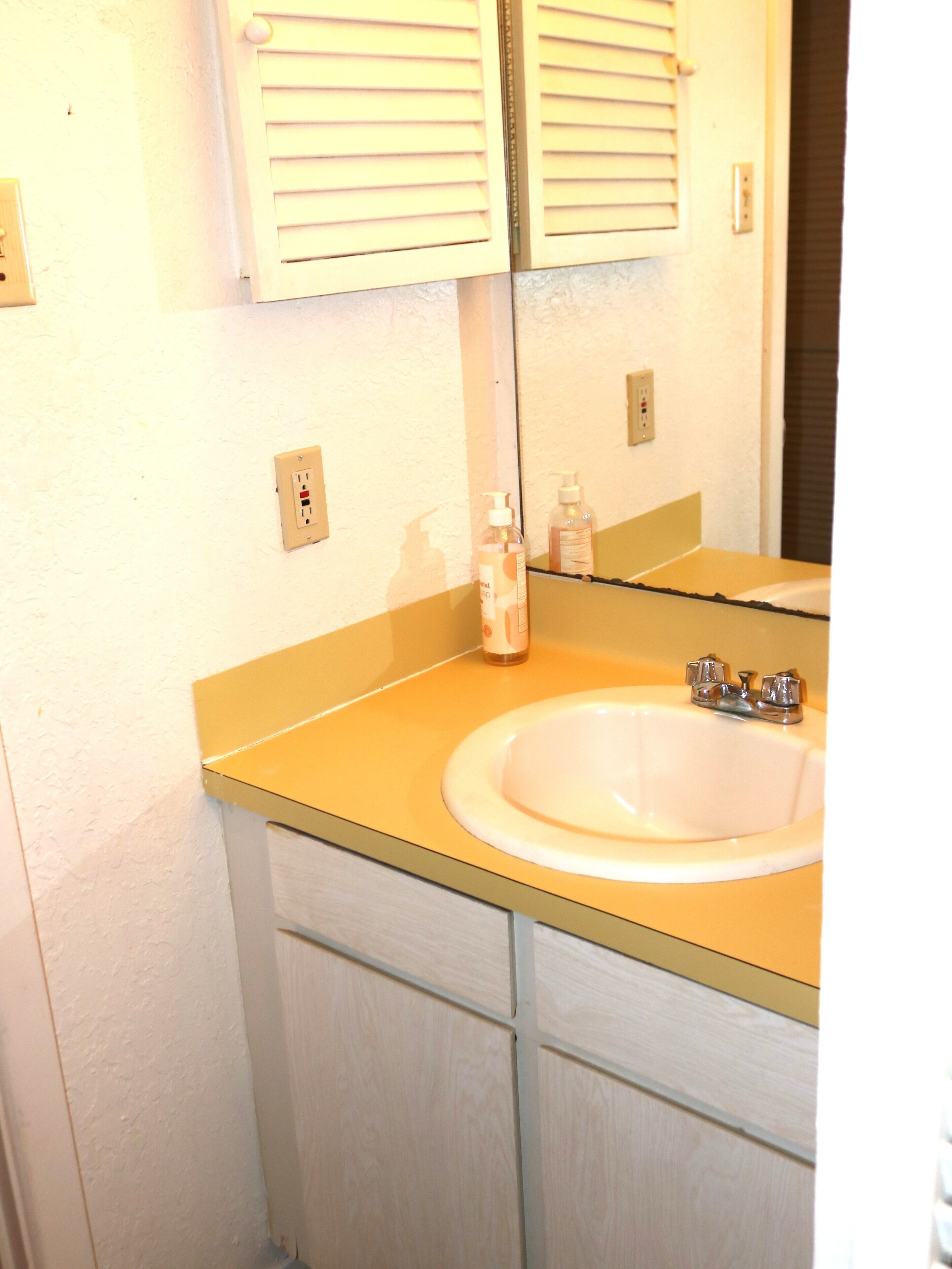 A bathroom with a white sink, yellow countertop, and a mirror above it. There are two bottles of hand soap on the counter.