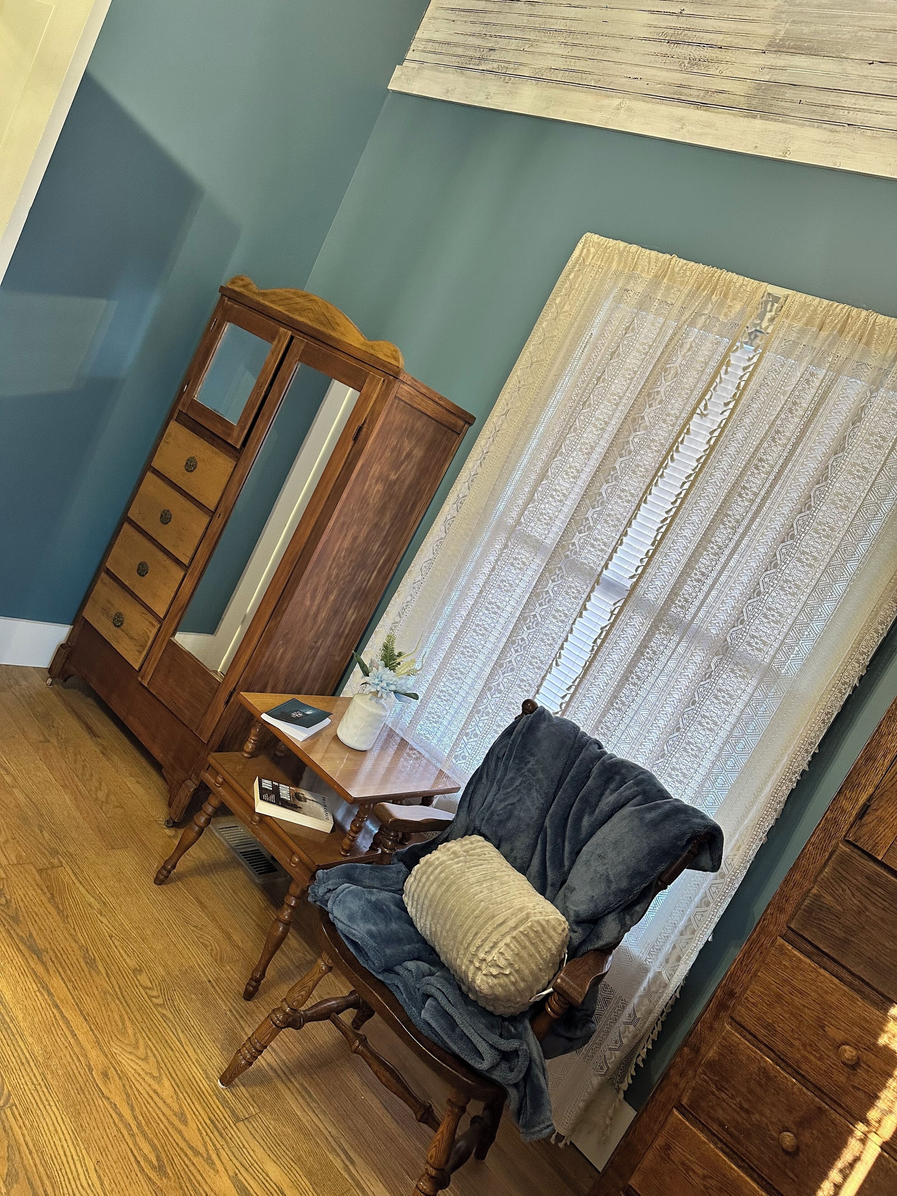 Una habitación con un aparador de madera, una silla con cojín, una mesita con un florero y una ventana con cortinas estampadas.