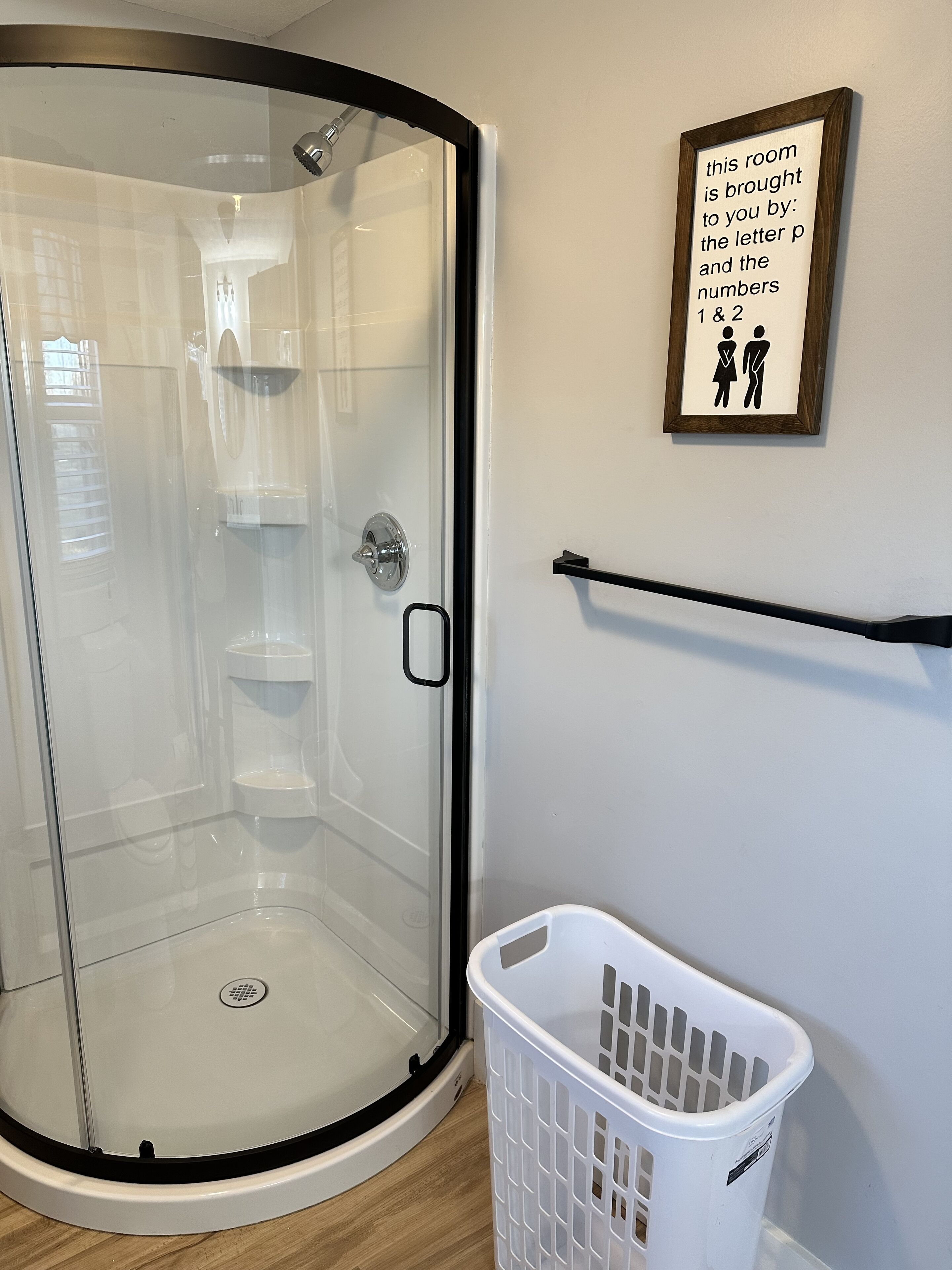 A shower room with a glass door, a framed sign on the wall, and a white laundry basket.