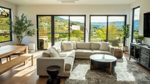A modern living room with a large sectional sofa, a round coffee table, and a dining area with a wooden table and bench. Large windows offer a view of a mountainous landscape. - Ultra Luxury Chelan Lake House w/ Sauna, Steam Room, Cold Plunge, and Hot Tub (Chelan)
