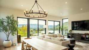 A spacious living room with a large sofa, a dining table with chairs, and a chandelier. There is a large window offering a view of the outdoors. - Ultra Luxury Chelan Lake House w/ Sauna, Steam Room, Cold Plunge, and Hot Tub (Chelan)