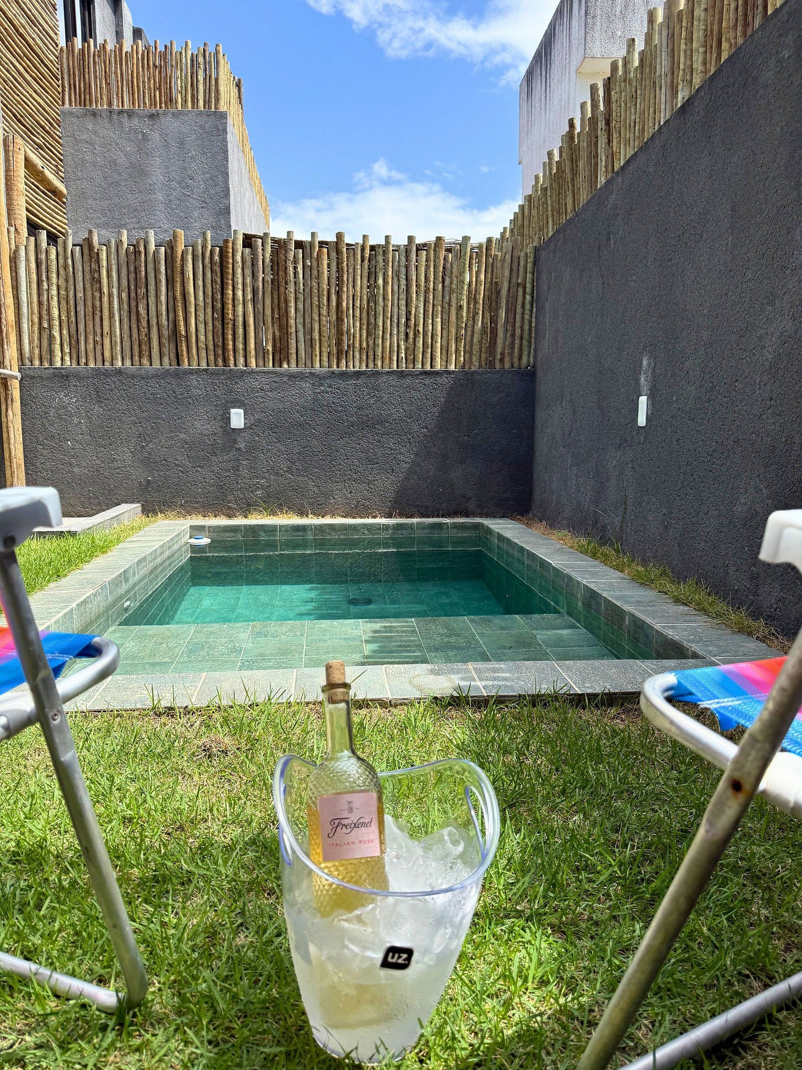 Una piscina con cerramiento de madera, una botella de champán y dos sillas de descanso.