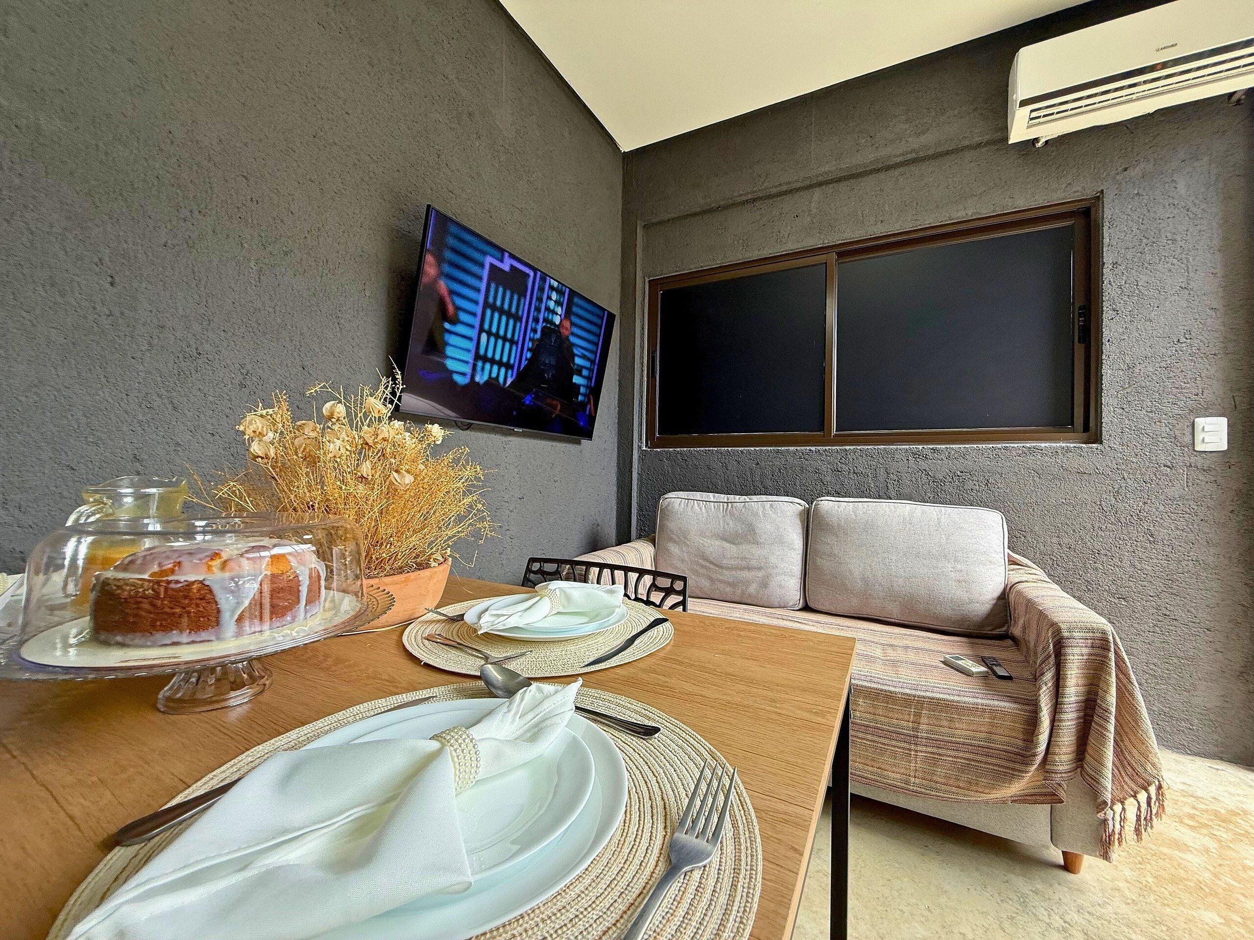 Uma sala de jantar com uma mesa posta para dois, uma televisão fixada na parede e um sofá com almofadas.