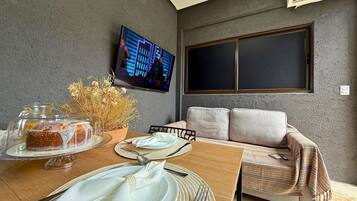 Uma sala de jantar com uma mesa posta para dois, uma televisão fixada na parede e um sofá com almofadas.