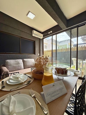 A dining area with a table set for two, a potted plant, a glass of orange juice, and a sign reading 'Bem vindos ao Studio Sra. Anna Lura & Esposa Grupo 05'. - MHV Milagres - Studio Guadalupe 05 (Passo de Camaragibe)