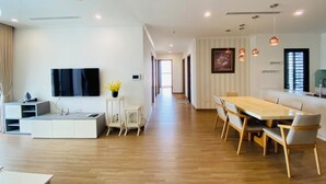 Ein modernes Wohnzimmer mit Flachbildfernseher, Essbereich mit Holztisch und -stühlen sowie Küchenzeile mit weißer Arbeitsplatte und Pendelleuchten.