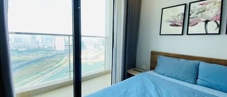 A bedroom with a large bed, a nightstand, and a view of a cityscape through a balcony.