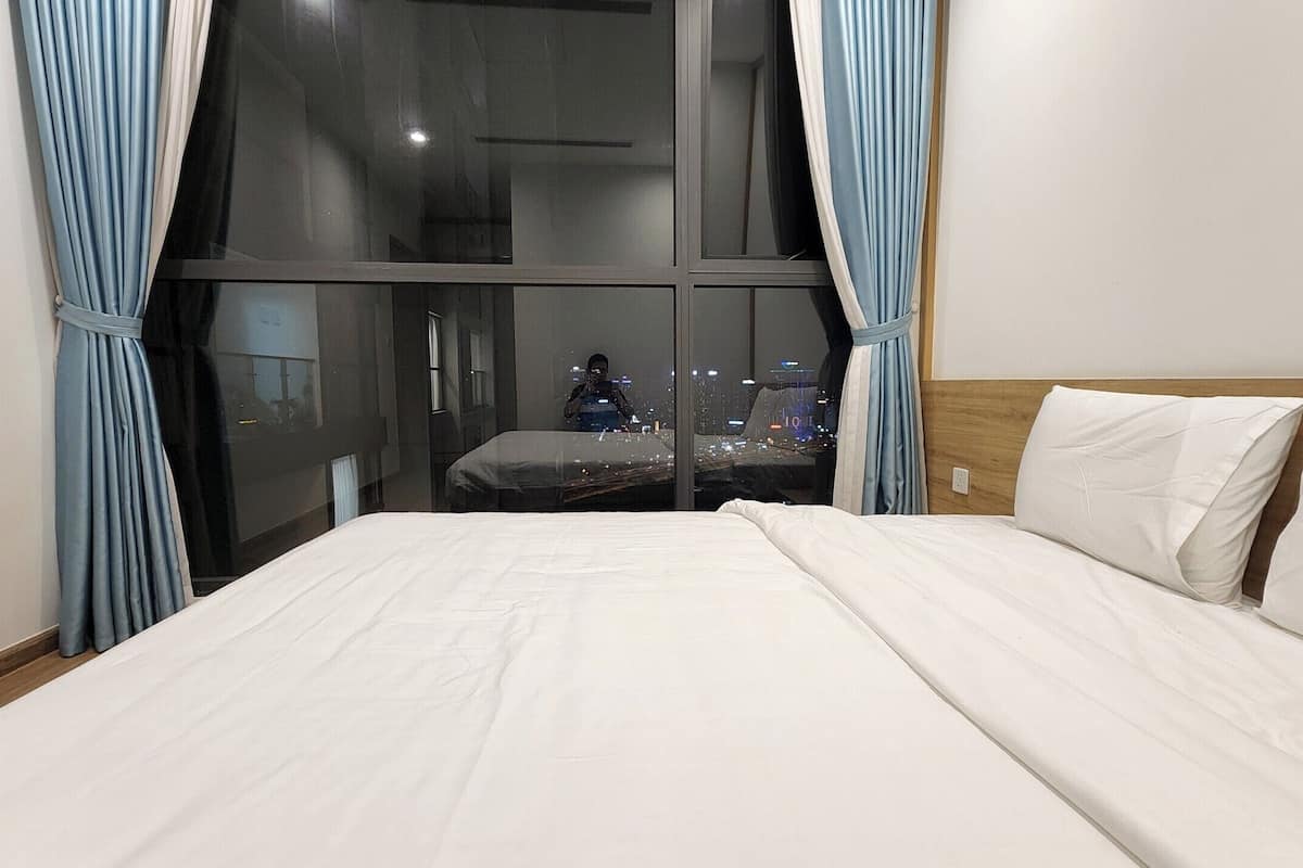 Une chambre d’hôtel avec un grand lit, une tête de lit en bois et une vue sur la ville grâce à de grandes fenêtres du sol au plafond.