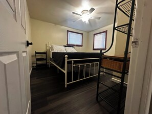 A compact bedroom with a single bed, a ceiling fan, two windows with blinds, a small chair, and a metal shelf unit. - Cozy 2 Bedroom in Fairbanks with Wifi (Fairbanks)