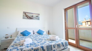 A bedroom with a bed, a painting, a nightstand, and a large sliding glass door to a balcony. - Villa Cycas by All4sardinia  (villasimius)
