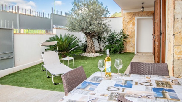 A patio with a table set for two, a bottle of wine, and a view of a tree and a stone wall. - Villa Cycas by All4sardinia  (villasimius)