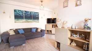 A modern living room with a grey sectional sofa, a flat-screen TV, wooden shelves with decorative items, and a large ocean-themed painting on the wall. - Villa Cycas by All4sardinia  (villasimius)