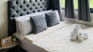 Een hotelkamer met een gestoffeerd hoofdeinde, een bed met wit en grijs beddengoed, een witte deken en een klein nachtkastje met een klok en een plant.