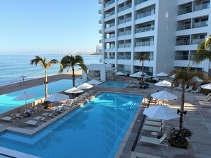 A pool area with lounge chairs and umbrellas, a beach, and a high-rise building in the background. - Exclusive Oasis with Spectacular Ocean View and VIP Amenities (Puerto Vallarta)