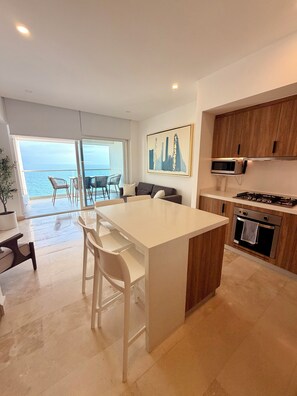 A modern kitchen with a central island, dining area with chairs, a view to the sea, and wooden cabinets.