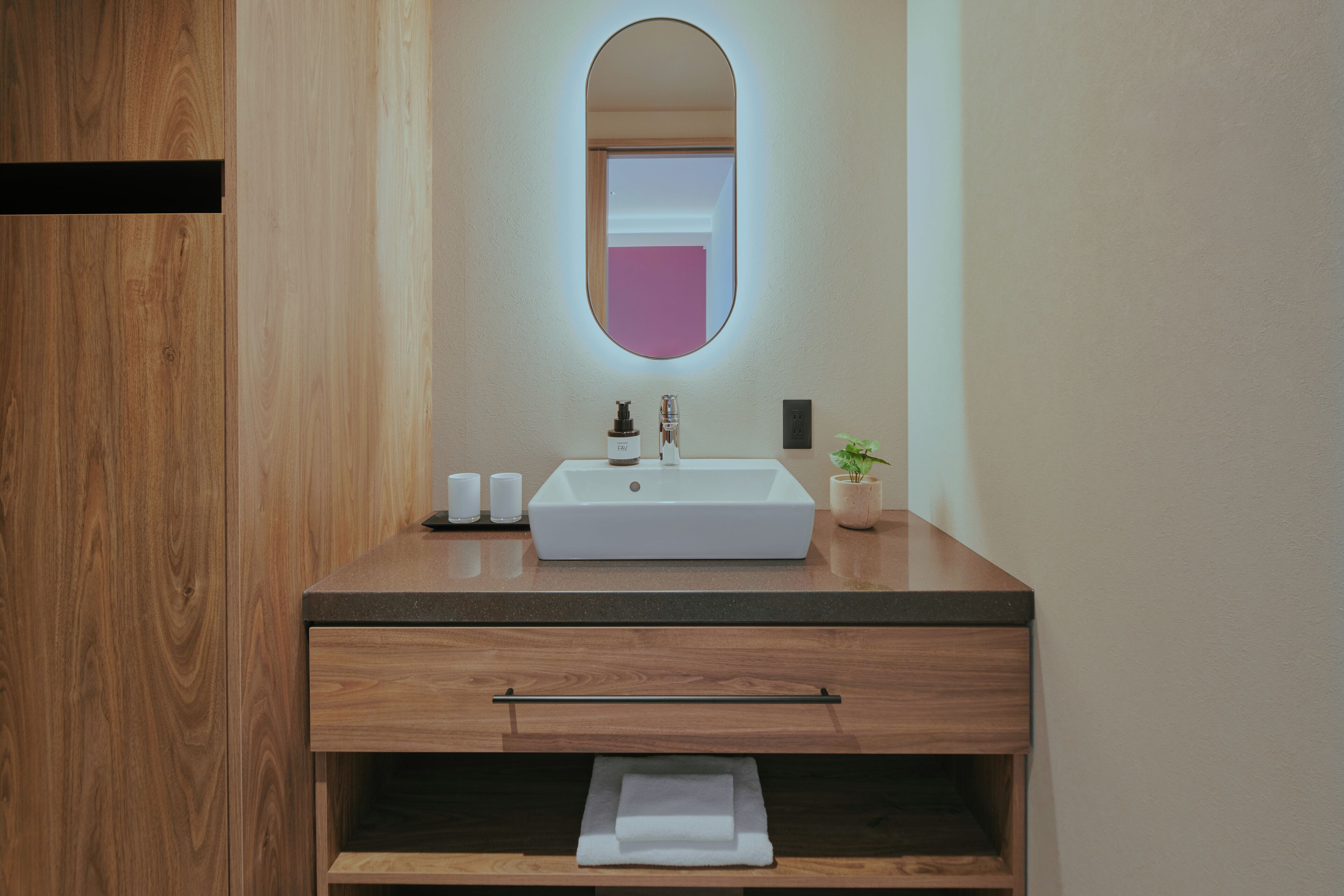 Une salle de bain moderne avec un meuble-lavabo en bois, un lavabo blanc, un miroir avec un cadre rose et une petite plante en pot.